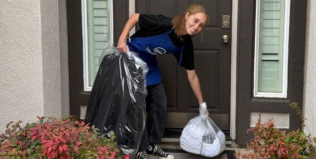 Man in apron inspects white shirt at a dry cleaner's, with a washing machine and hanging clothes visible.
