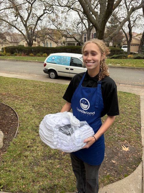 Woman in blue apron holding a stack of white items, smiling. White van in background.