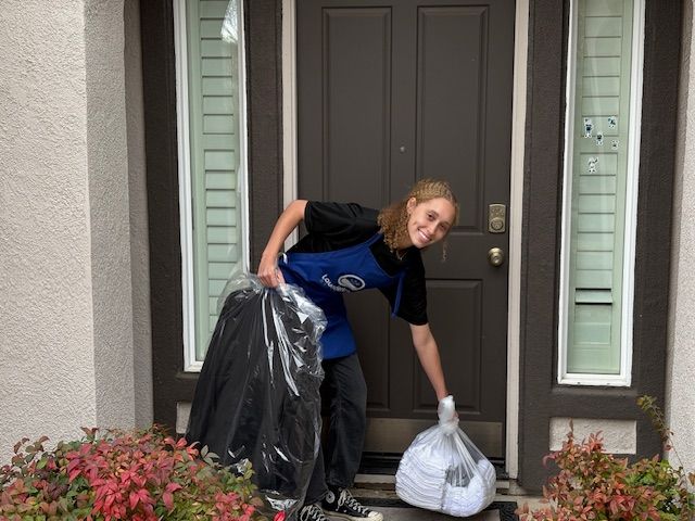 Person delivering bags on a doorstep. Holding a black trash bag and a white bag, smiling.
