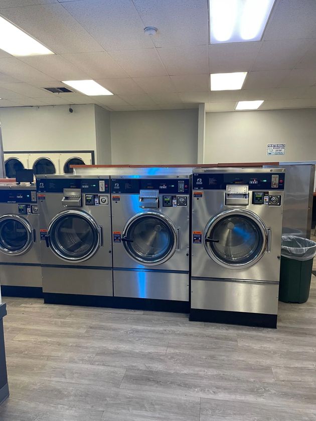 Row of stainless steel washing machines in a laundromat. Fluorescent lights and a trash can are visible.