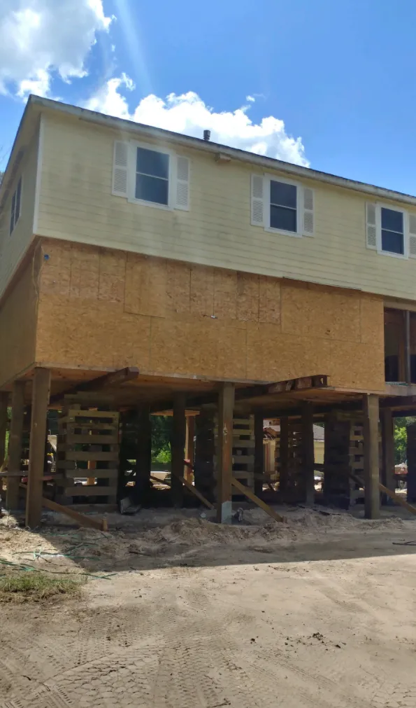 Elevated house under construction on tall wooden supports, with unfinished lower walls and sandy ground below.