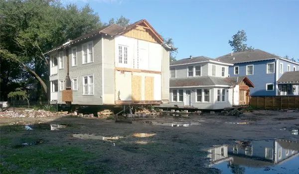 Two houses under construction beside muddy ground and puddles, with trees in the background.