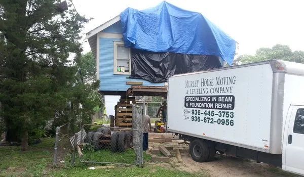 House being lifted by a crane beside a white utility truck, with a blue tarp covering the roof.