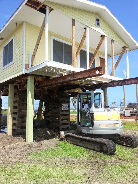 House lifted on stilts with a tracked excavator underneath, showing structural support work in progress.