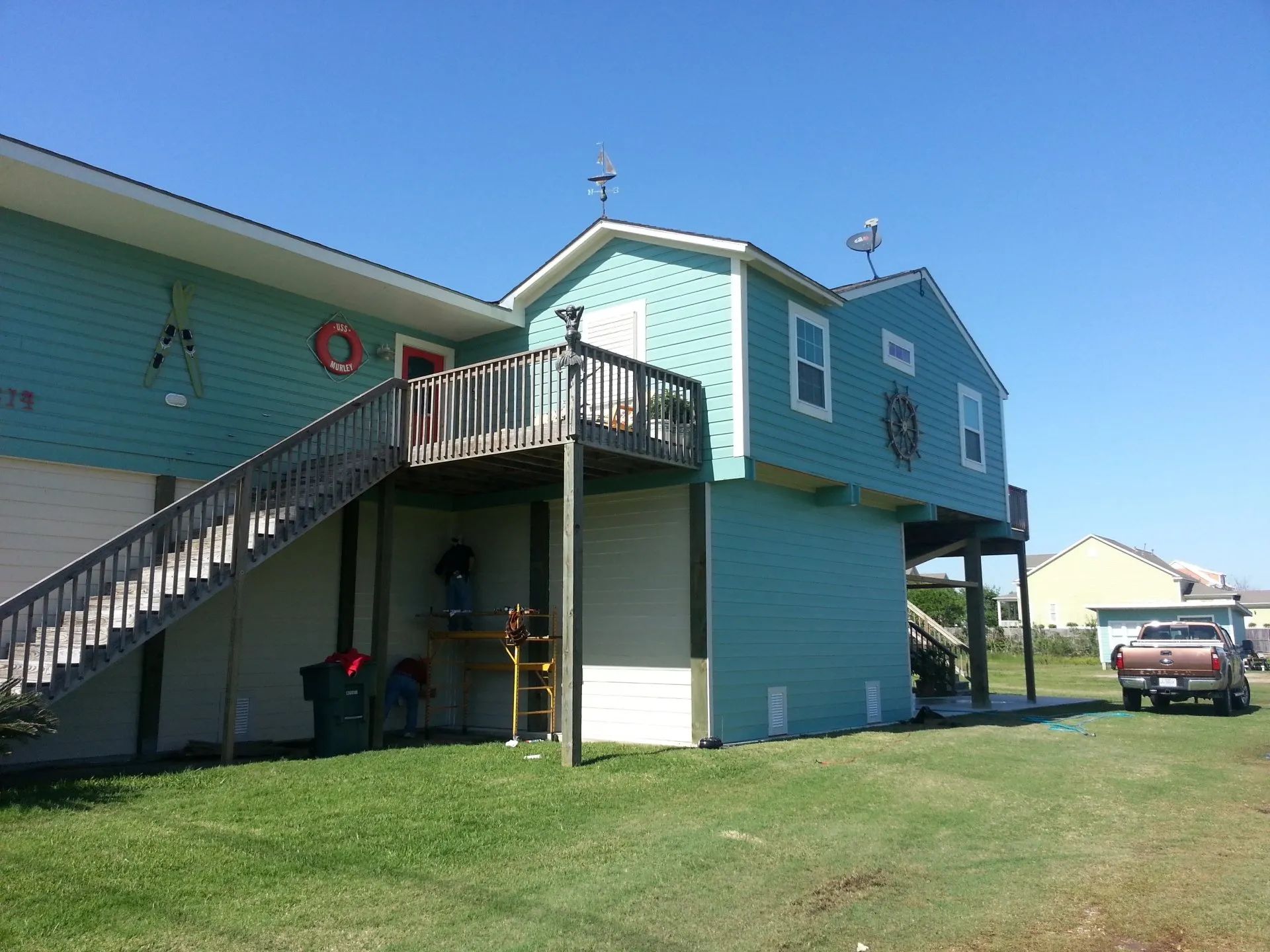 Blue elevated house with exterior stairs and deck on a grassy lot under a clear sky