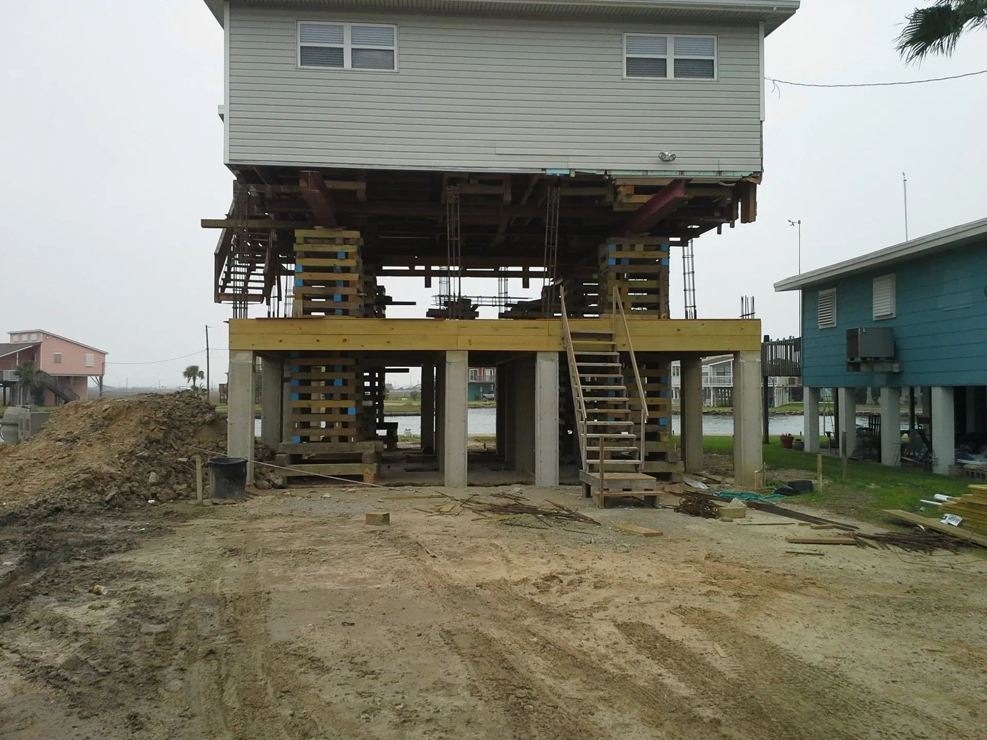 Elevated house on stilts with stairs on a dirt lot under construction, beside a blue house
