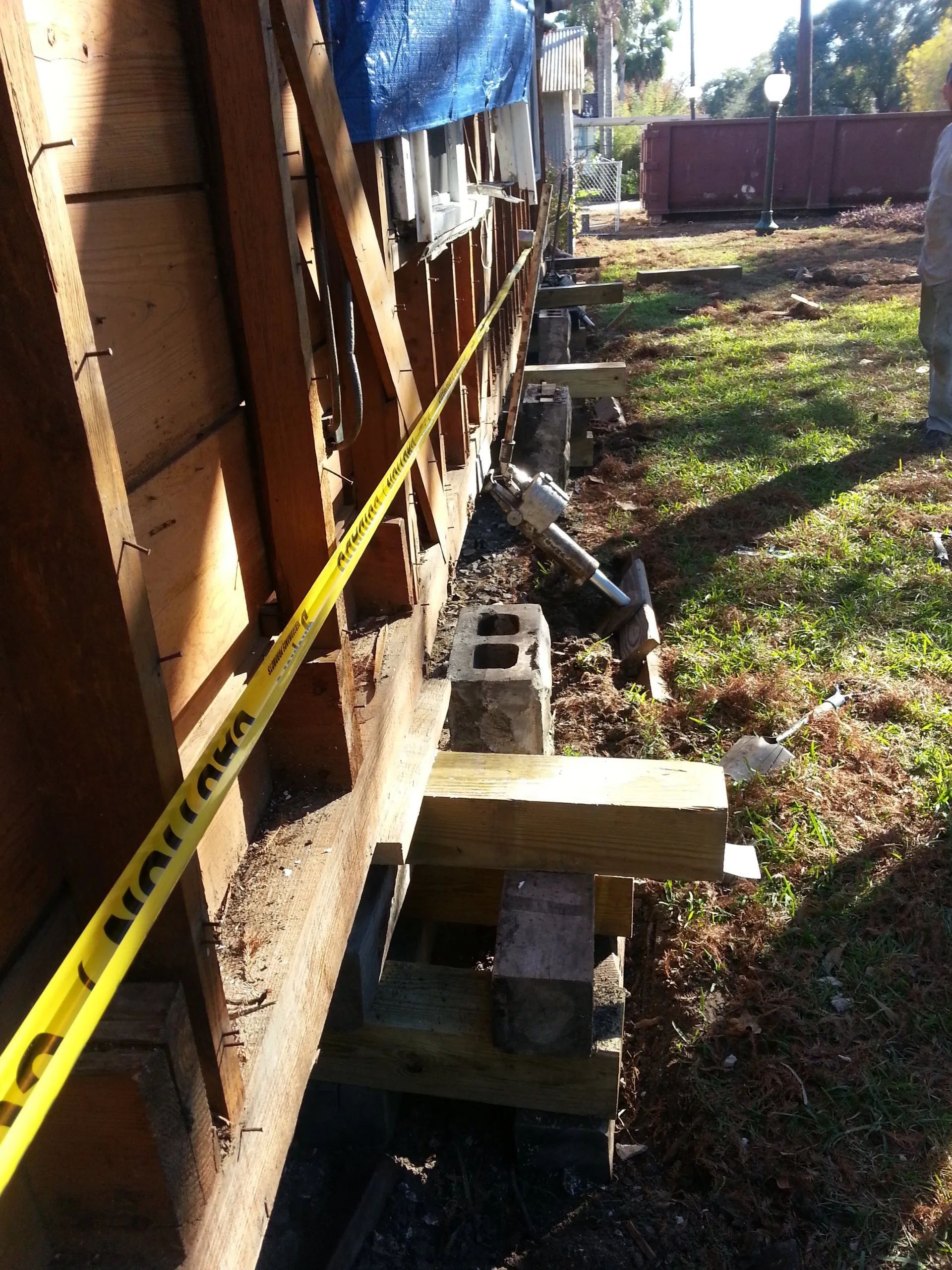 Side view of a house foundation repair with yellow caution tape, cinder blocks, and wooden supports along the wall