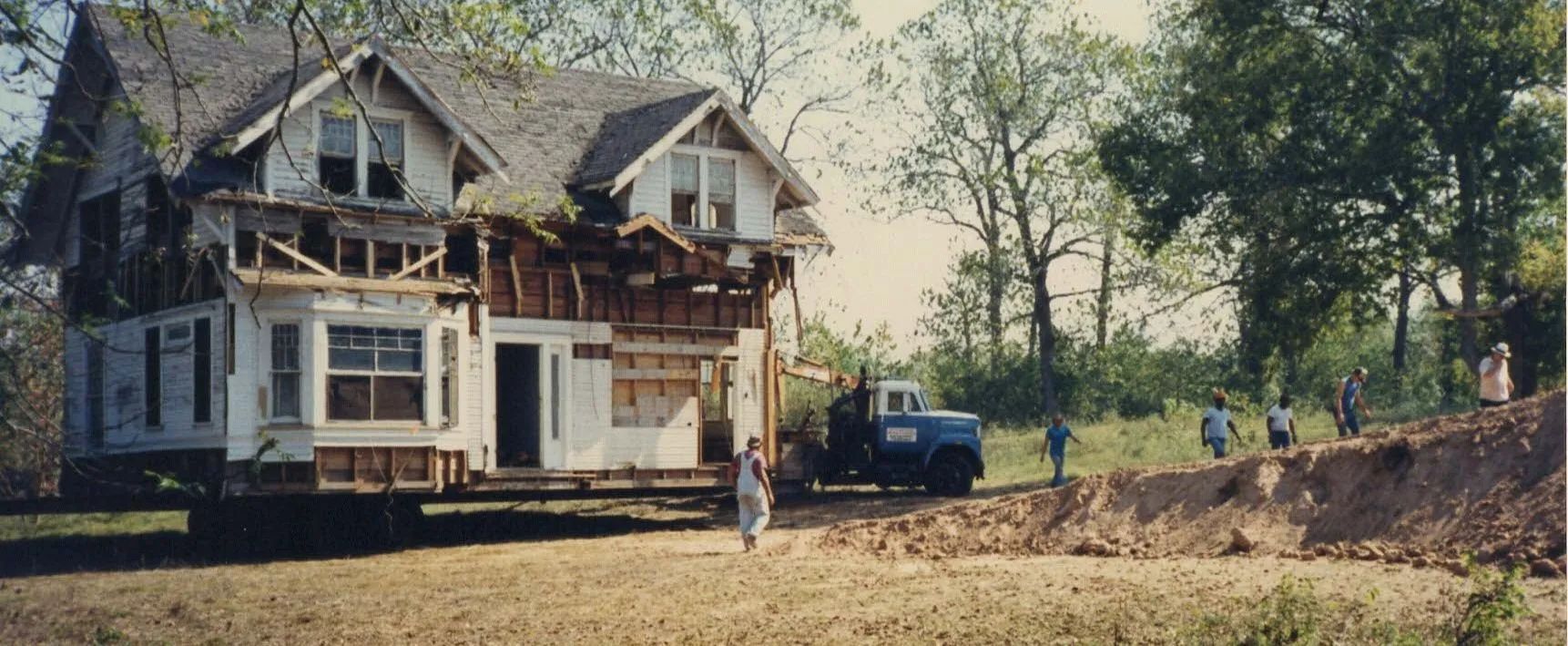 Old white house under renovation beside a dirt lot, with scaffolding and trees nearby.