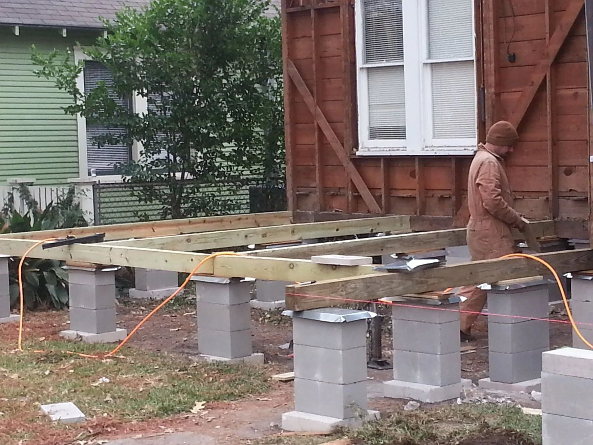 Worker building a raised wooden platform on concrete blocks beside a house