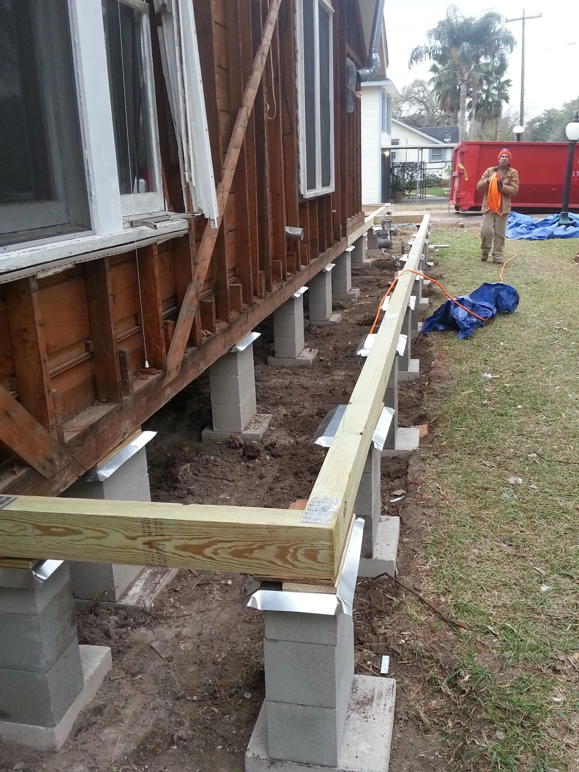 Elevated house under construction on concrete piers, with wooden framing and workers in the yard.