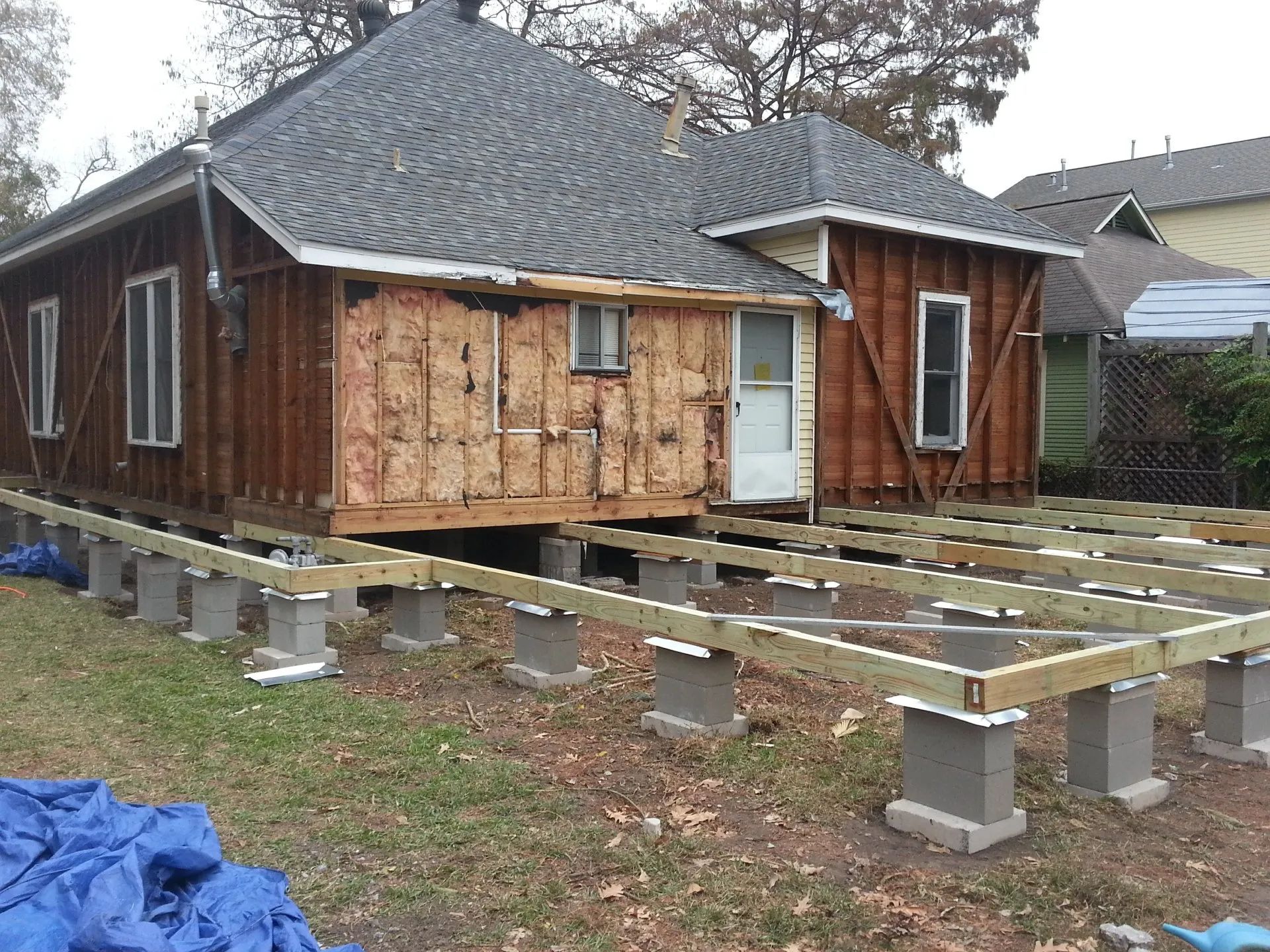 Wood house raised on concrete piers during foundation repair, with exposed joists and blue tarp in yard.