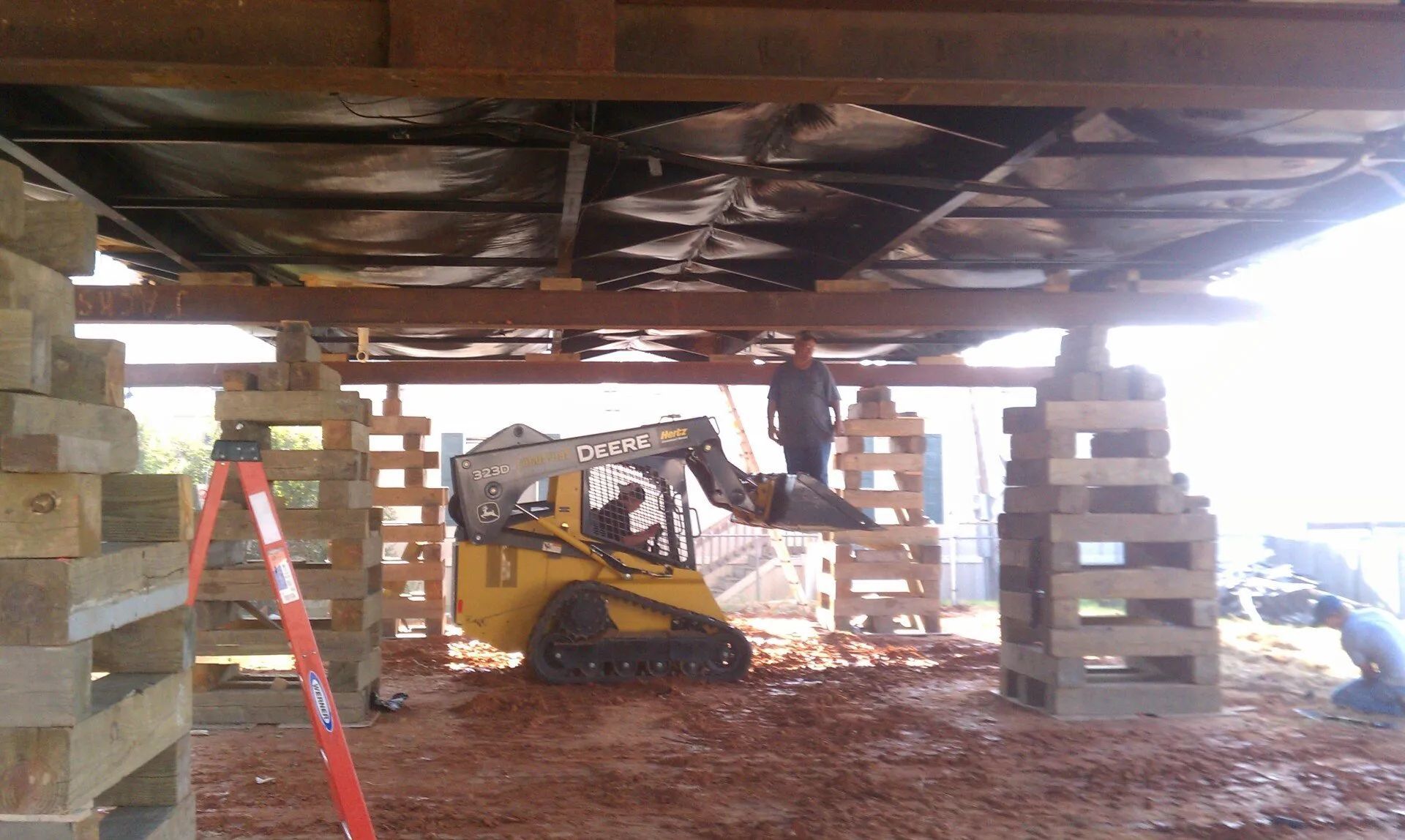 Small yellow excavator under a raised building, supported by stacked cinder blocks and a red ladder nearby.