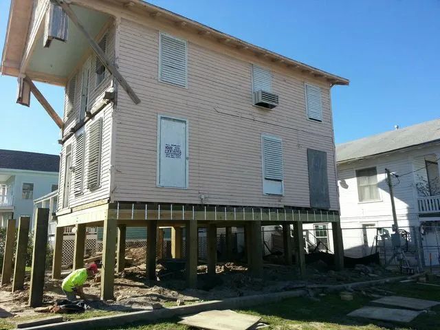 Raised house on stilts with unfinished lower level and a worker crouching outside in a yard