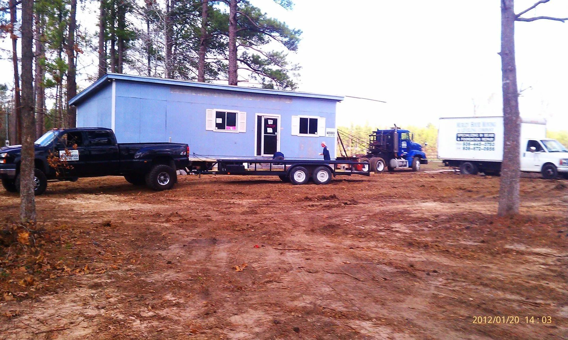Black pickup and blue mobile home on a dirt lot among tall pine trees.