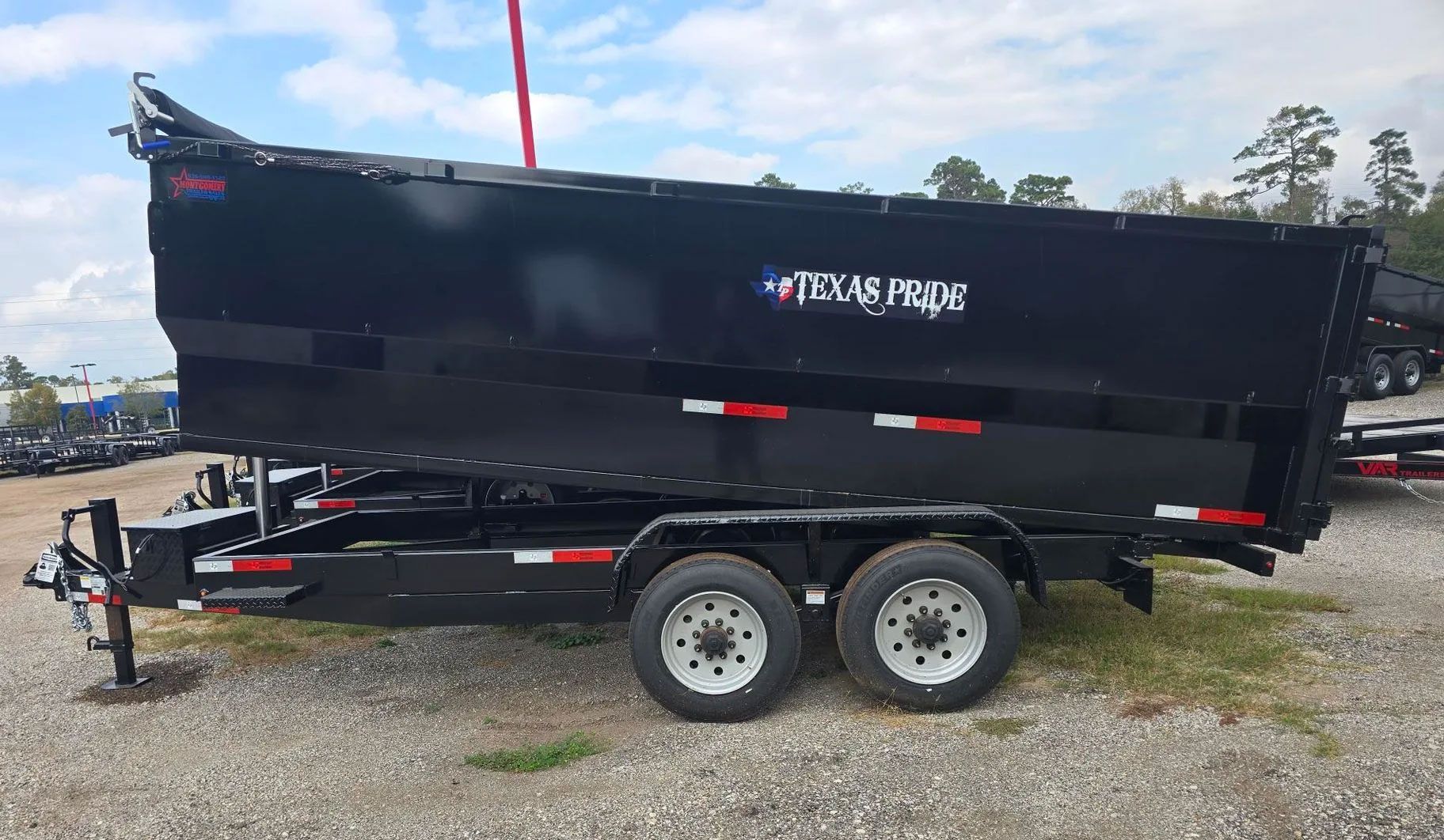Black Texas Pride dump trailer on gravel lot, side view, with dual axles and raised front