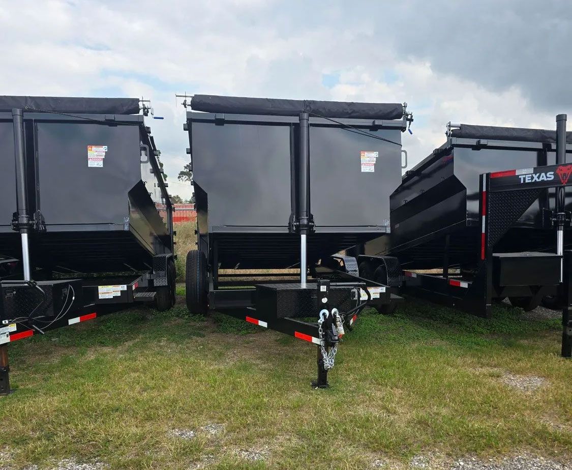 Three black dump trailers parked on grass, viewed from behind under a cloudy sky