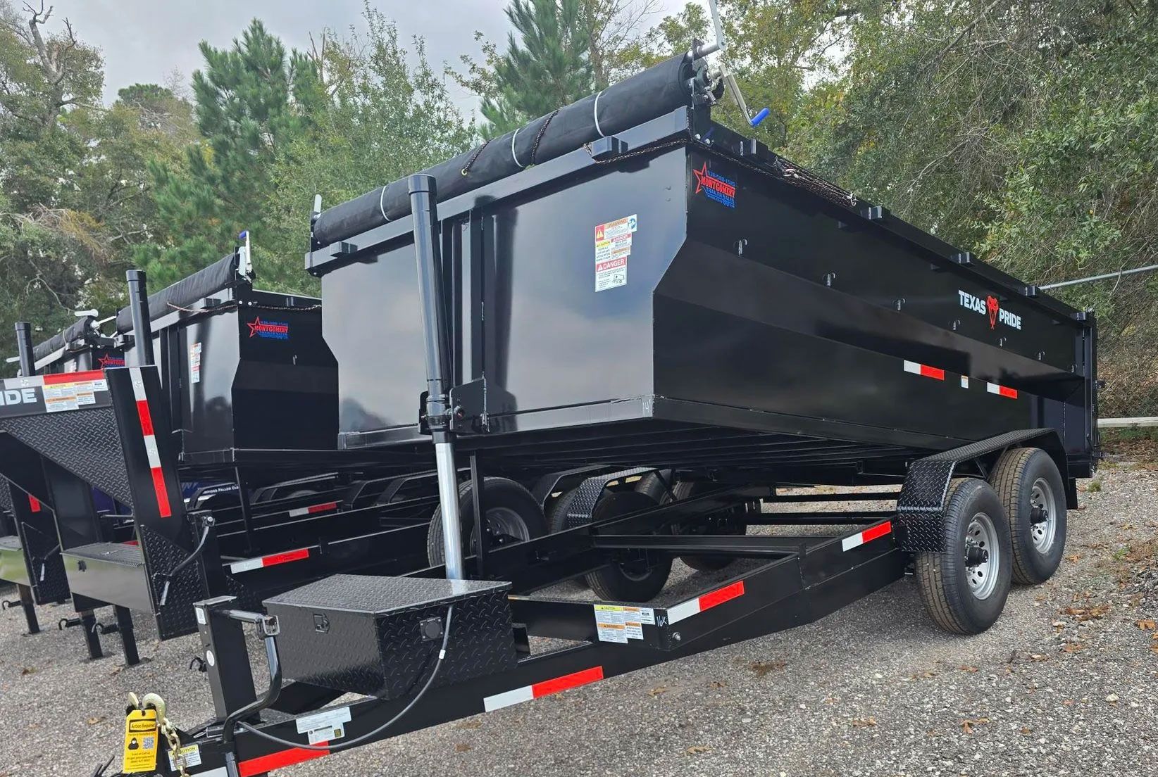 Black dump trailer parked on gravel, with another trailer behind it and trees in the background.