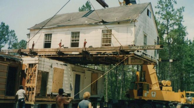 House being lifted by crane during relocation, with workers and scaffolding visible underneath