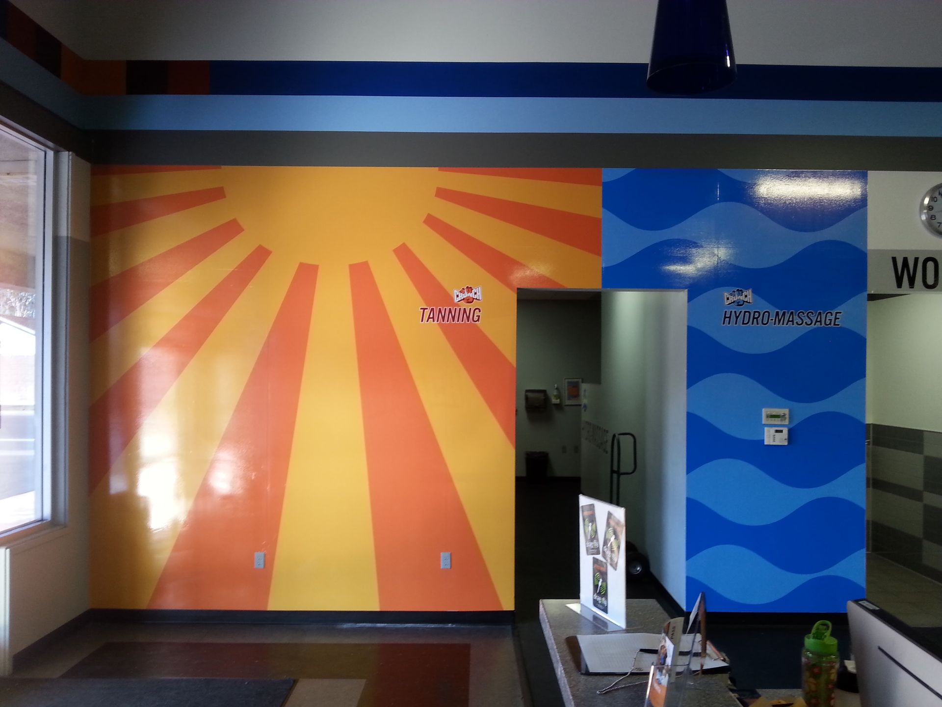 Colorful mural with orange sun rays and blue waves in a hallway with doors and a wall-mounted display
