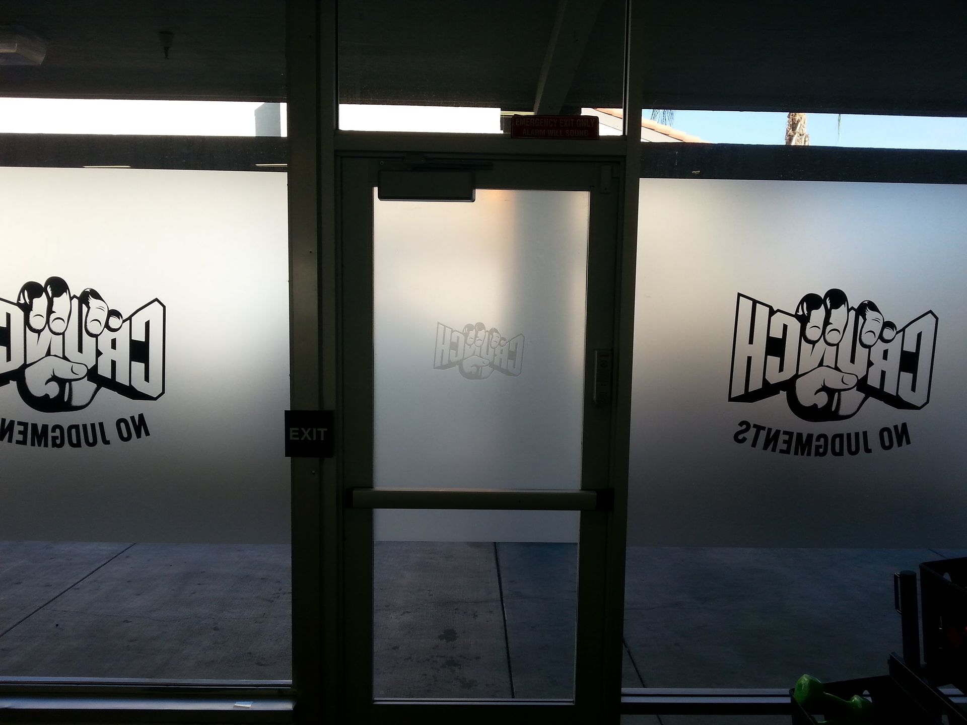 Inside view of a glass storefront with mirrored logos on the doors and a bright sidewalk outside.