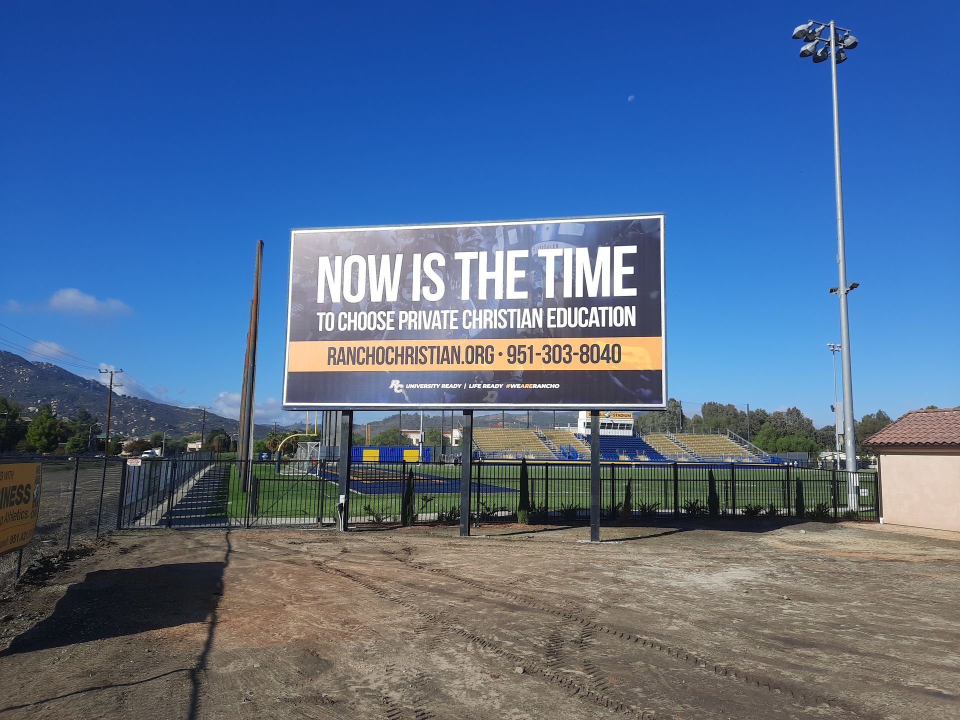 Billboard in a fenced field reads “NOW IS THE TIME” under a clear blue sky