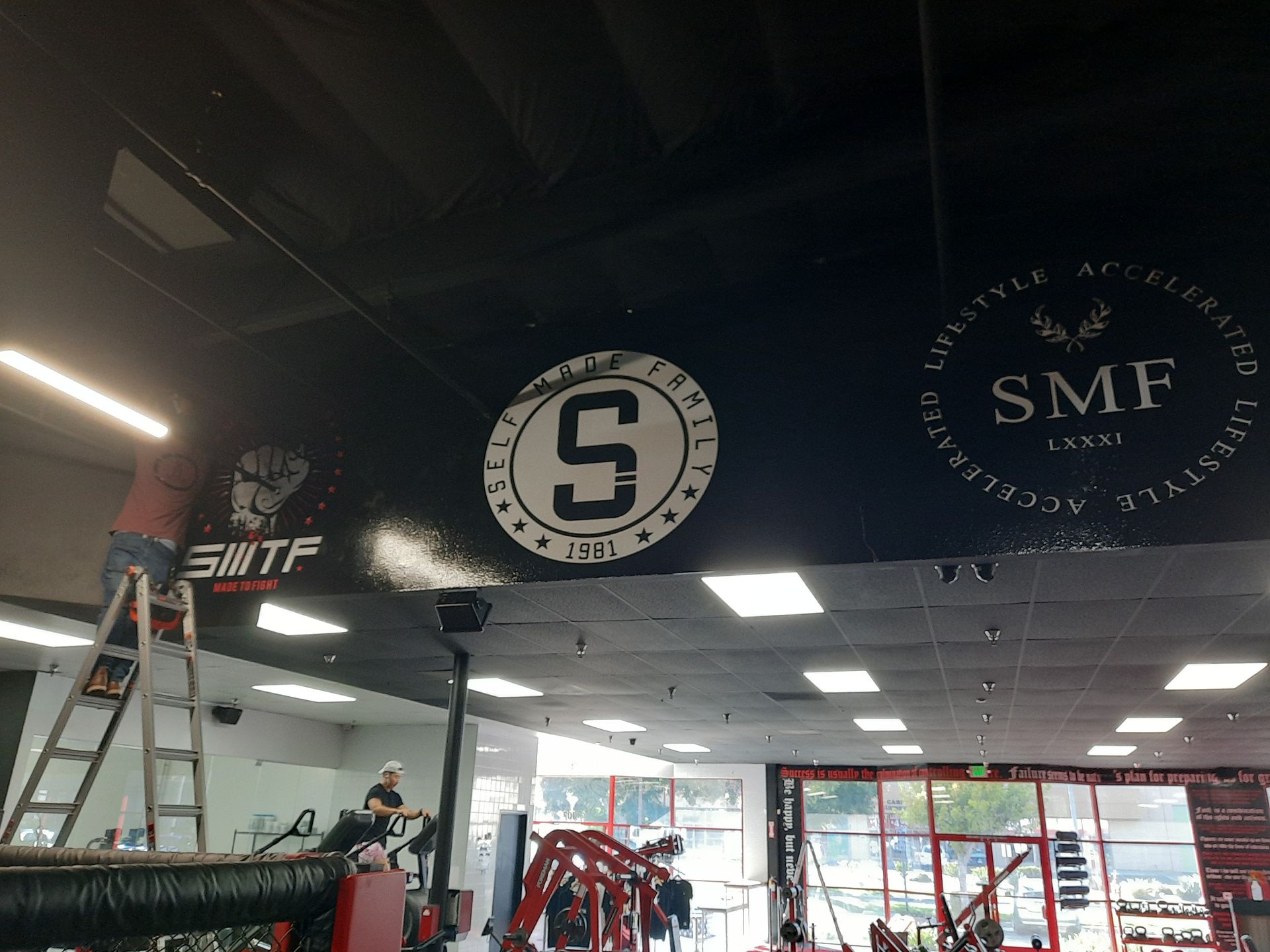 Gym interior with red equipment, mirrored walls, and large black SMF logos on the ceiling