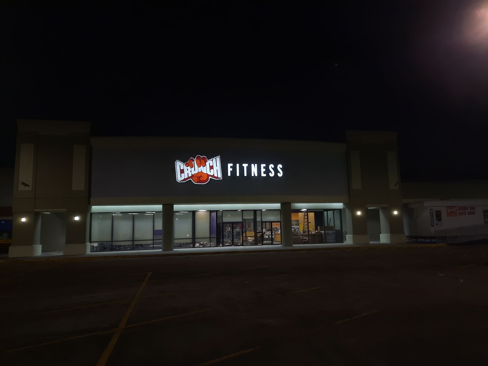 Nighttime exterior of a lit fitness gym storefront in a shopping center parking lot