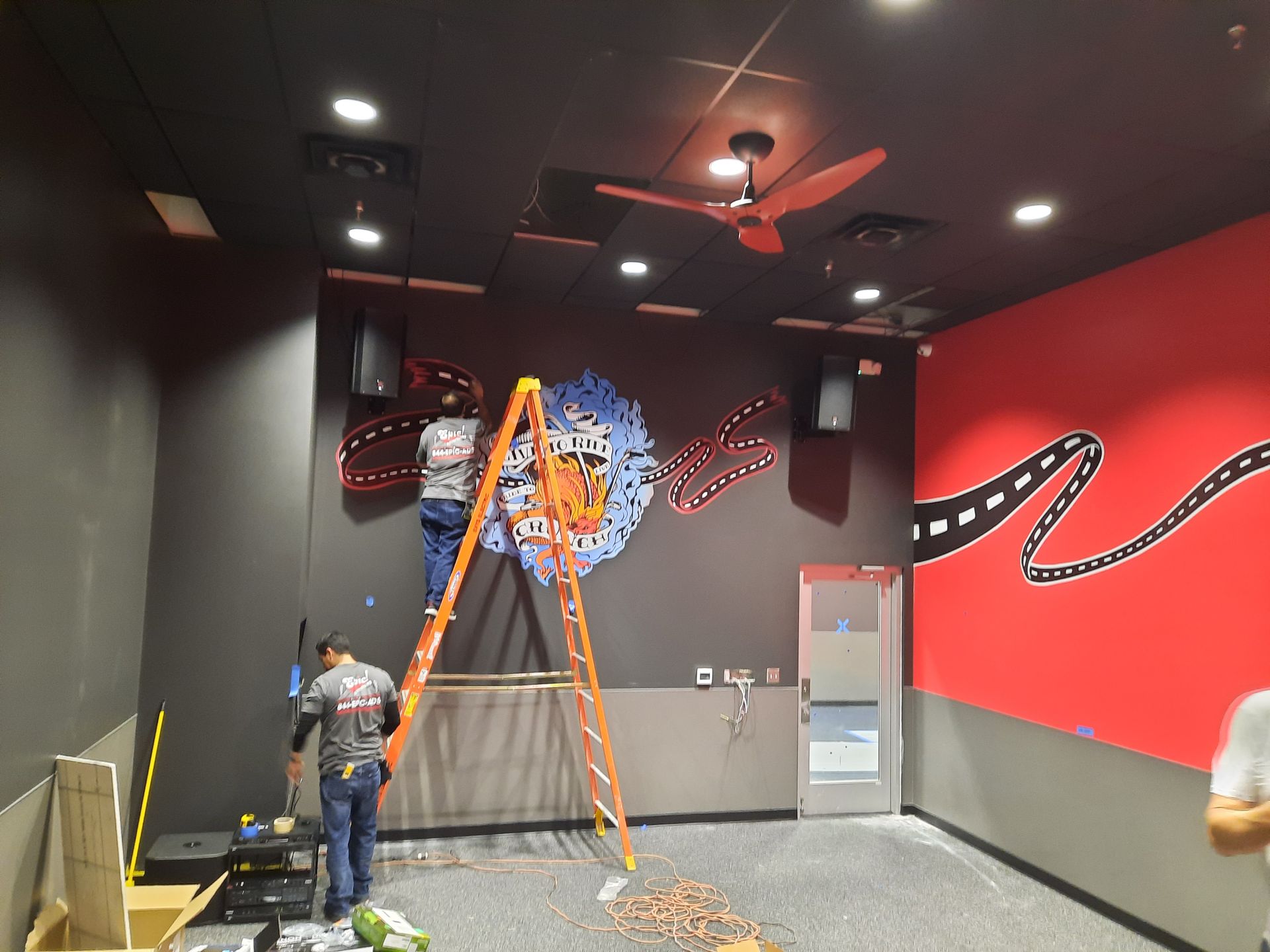 Worker on ladder painting a black-and-red room with movie-themed wall art and ceiling lights.