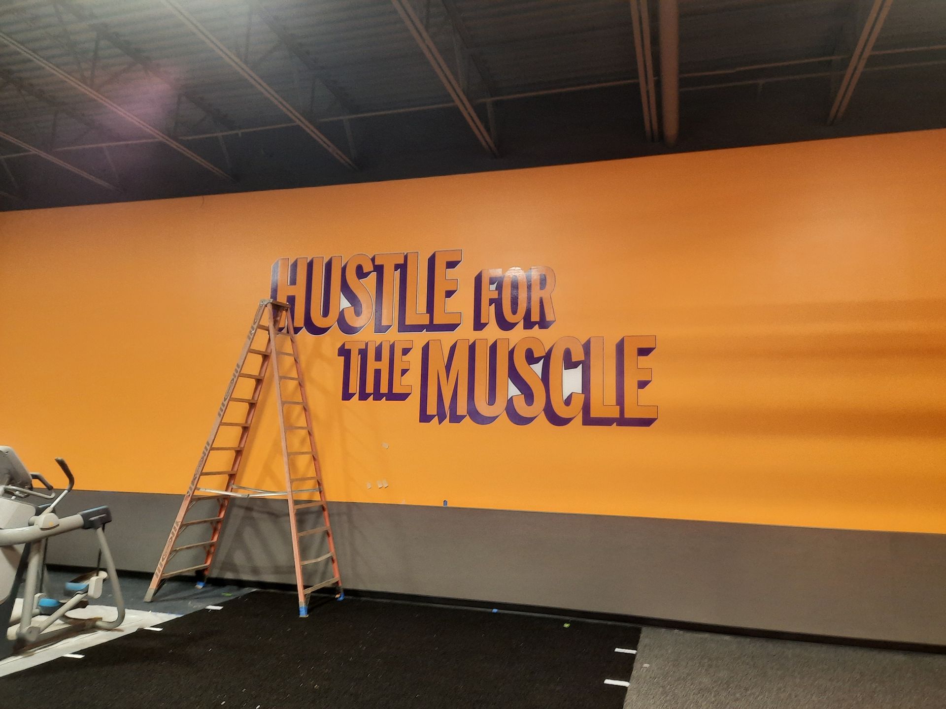 Gym wall mural reading “Hustle for the Muscle” beside a ladder in a fitness room