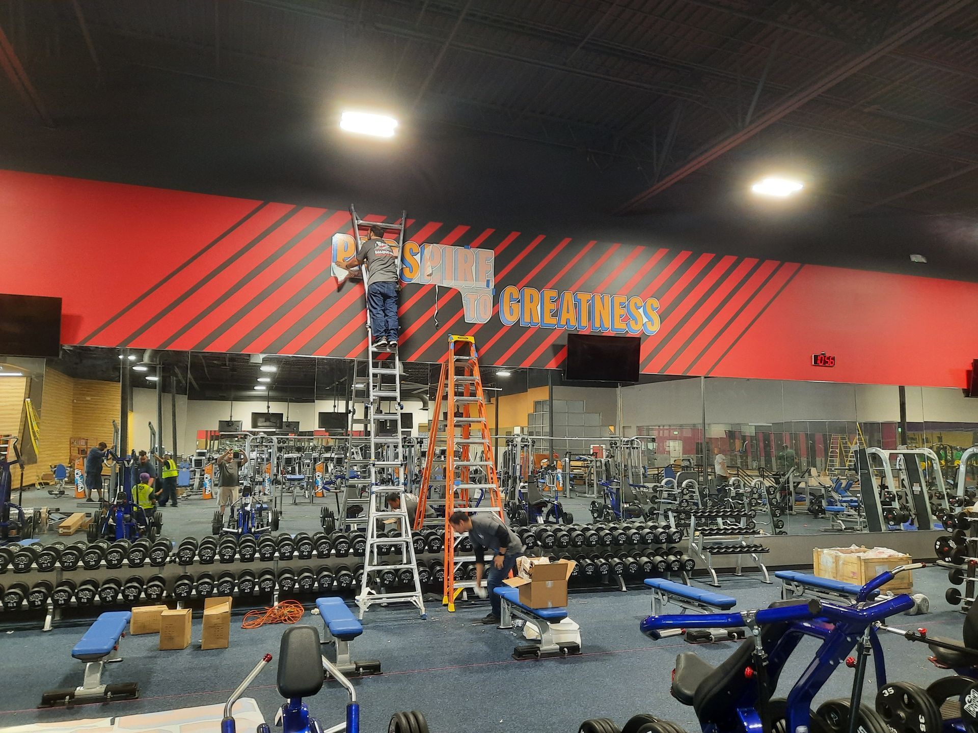 Gym interior with mirrors, weights, red wall, and a person on an orange ladder