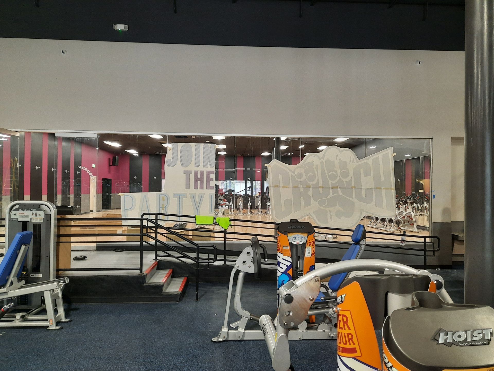 Gym interior with equipment, benches, and mirrors; orange and gray machine in the foreground.