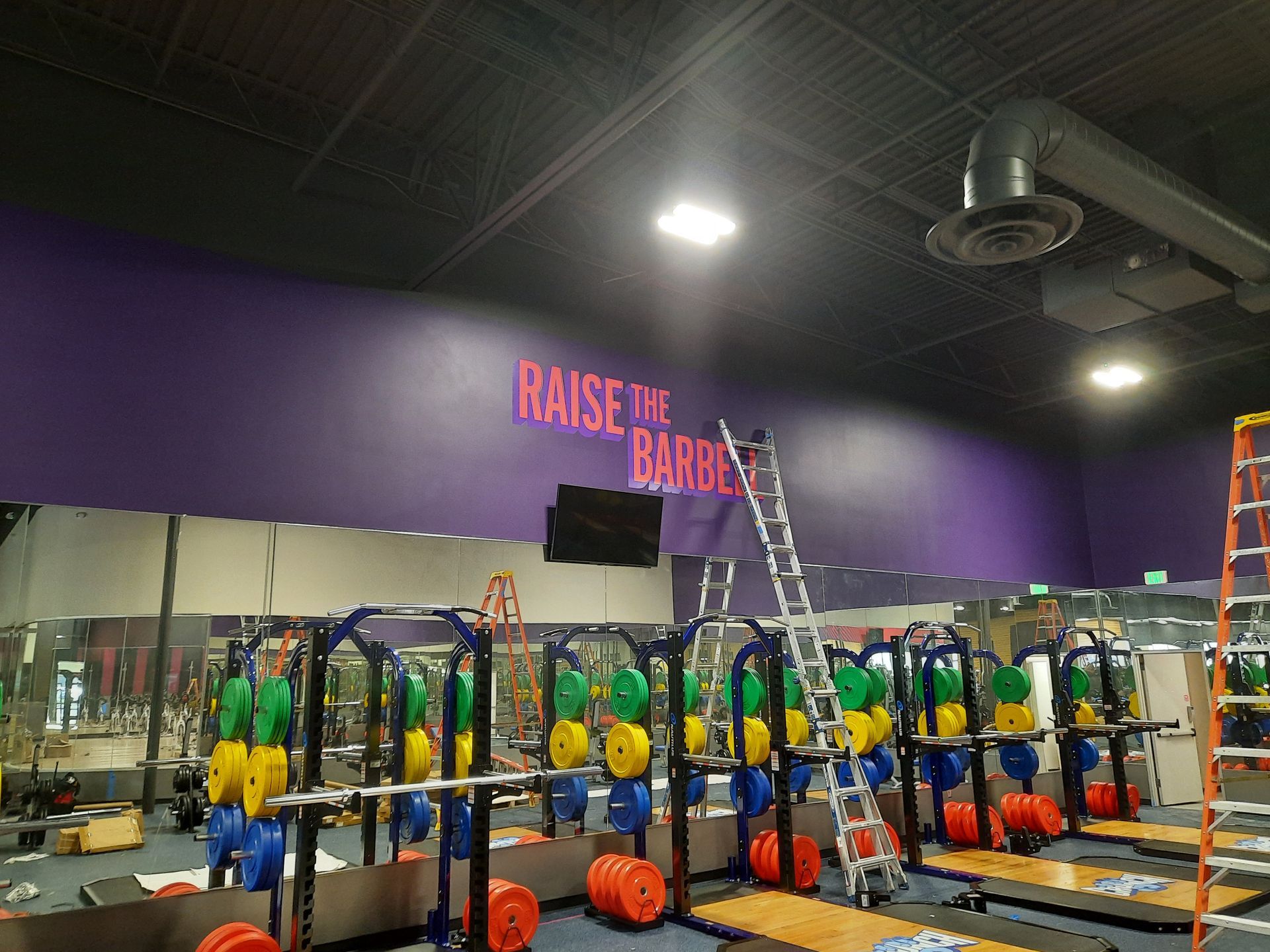 Gym interior with racks of yellow and green kettlebells, a ladder, and neon wall lettering reading “RAISE THE BAR.”
