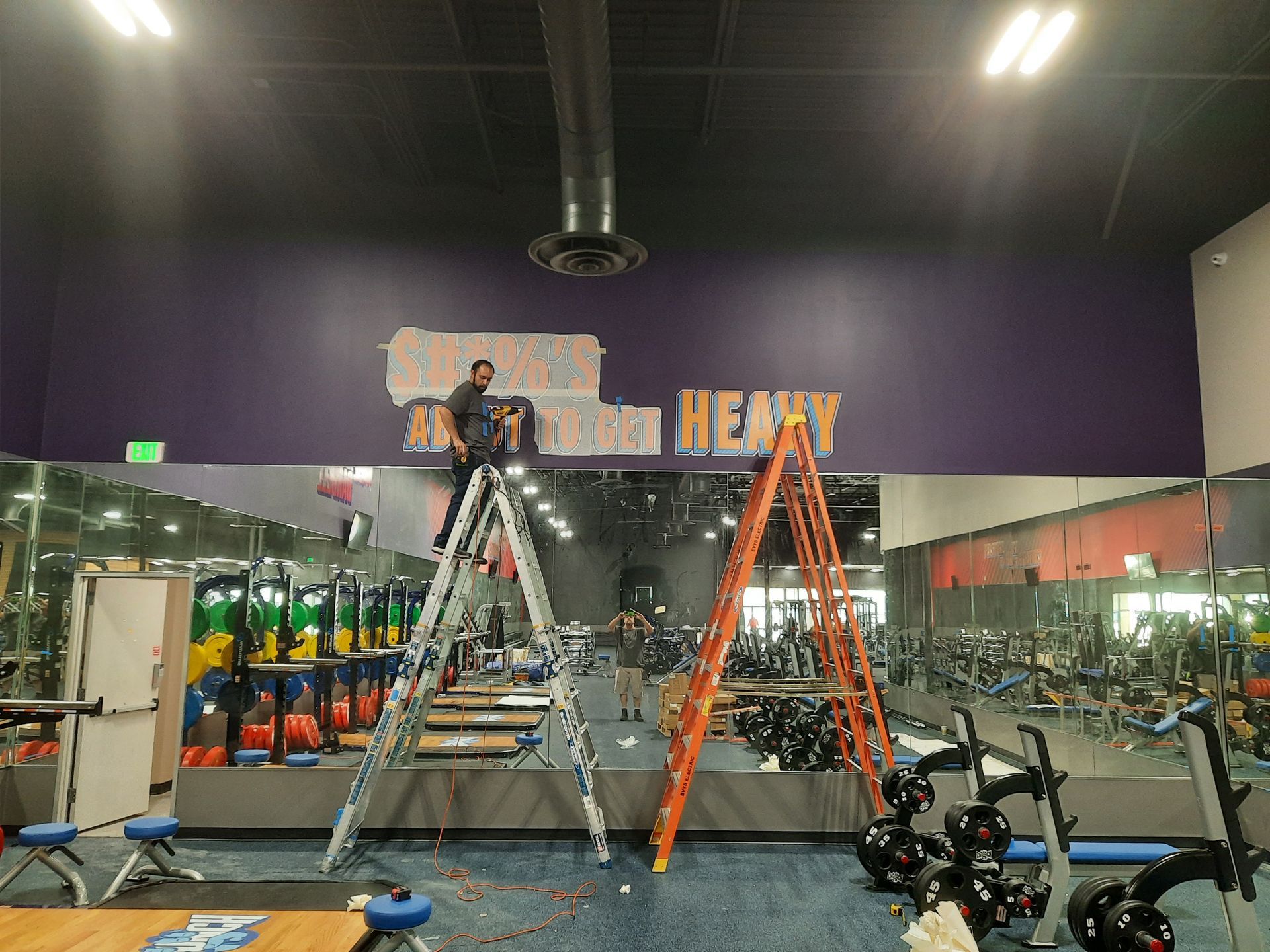 Two workers on ladders at a gym, installing overhead signage above mirrors and exercise bikes.