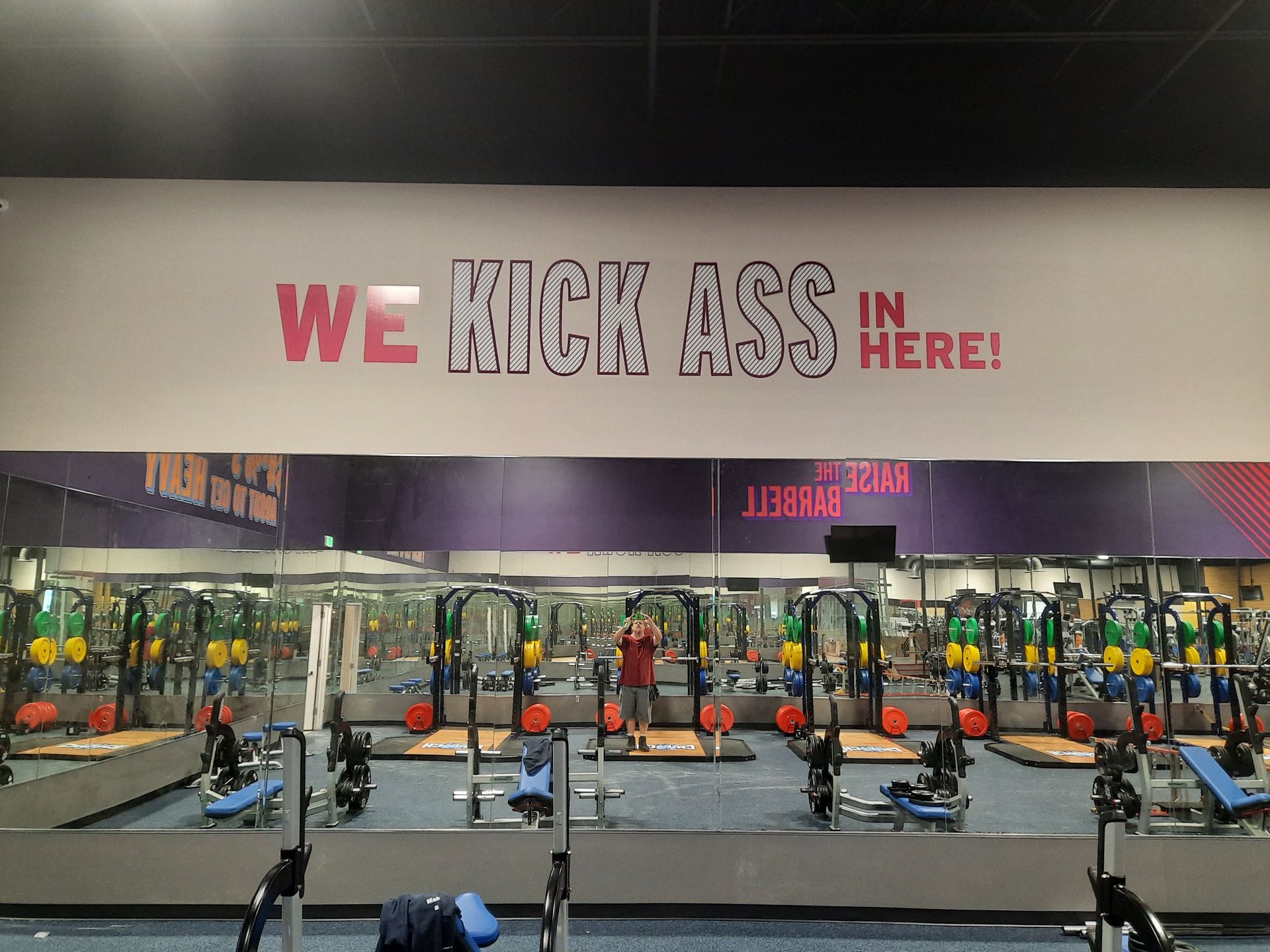 Gym with rows of weight benches and red signs reading “WE KICK ASS IN HERE!”