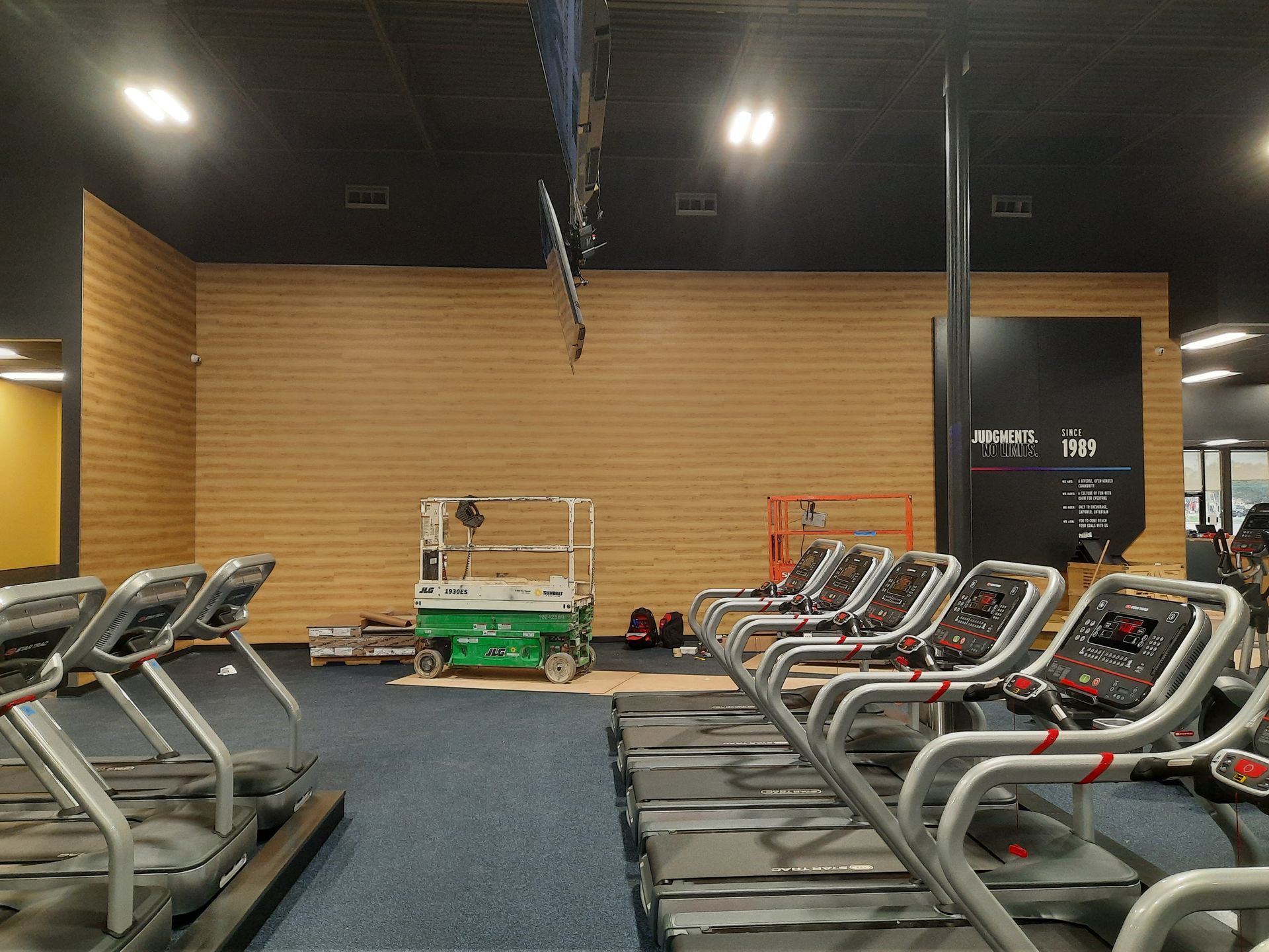 Empty gym cardio area with rows of treadmills facing a wooden wall under dim lighting