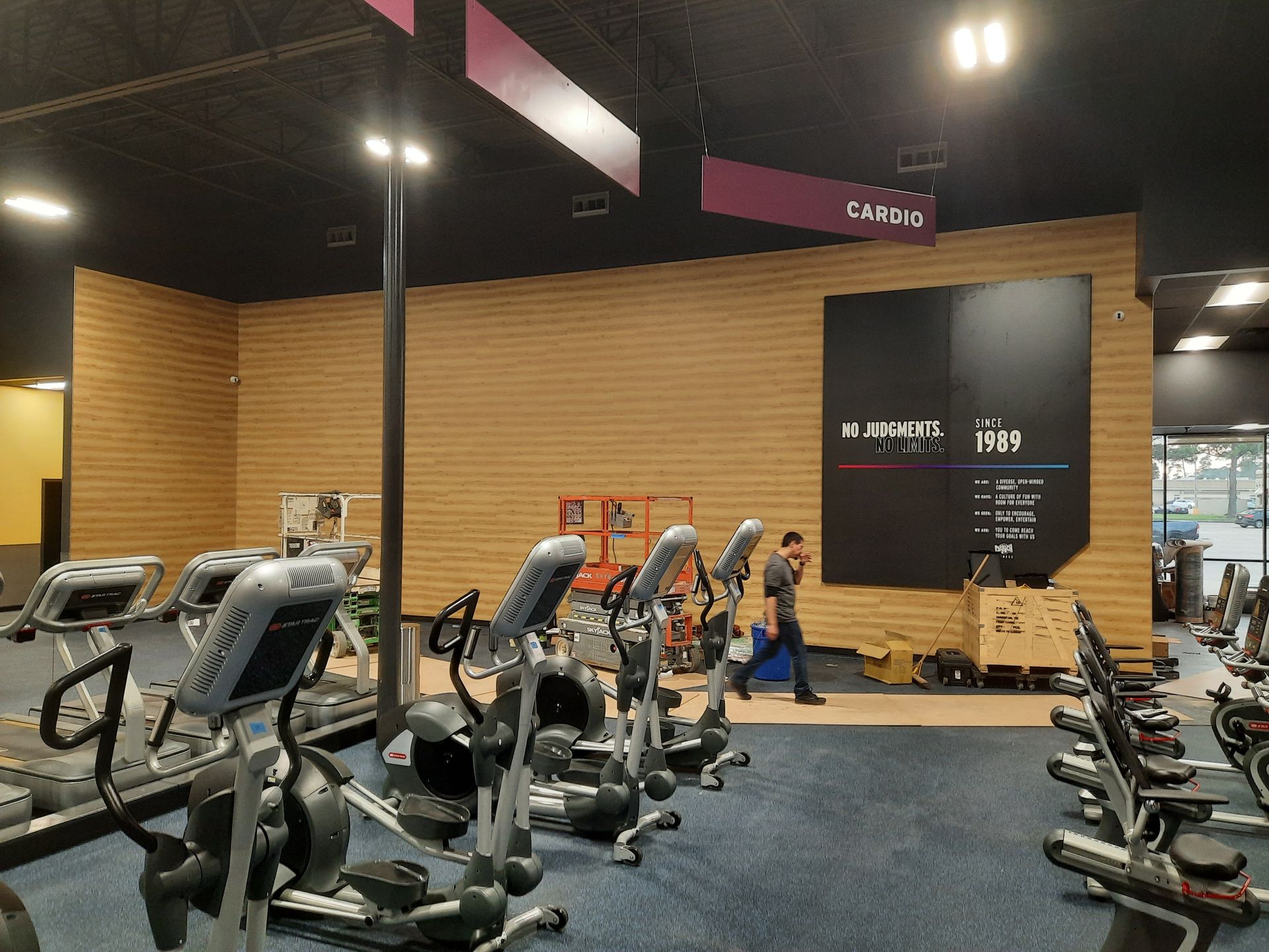 Gym interior with rows of exercise bikes and dumbbells on a blue floor, wood-paneled walls behind.