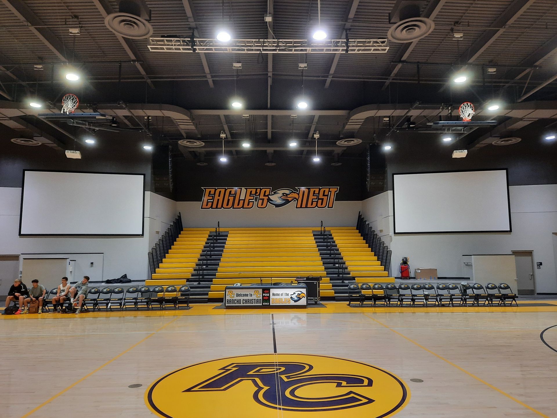 Empty indoor gym with bleachers, stage, and scoreboard walls above a yellow center-court logo.