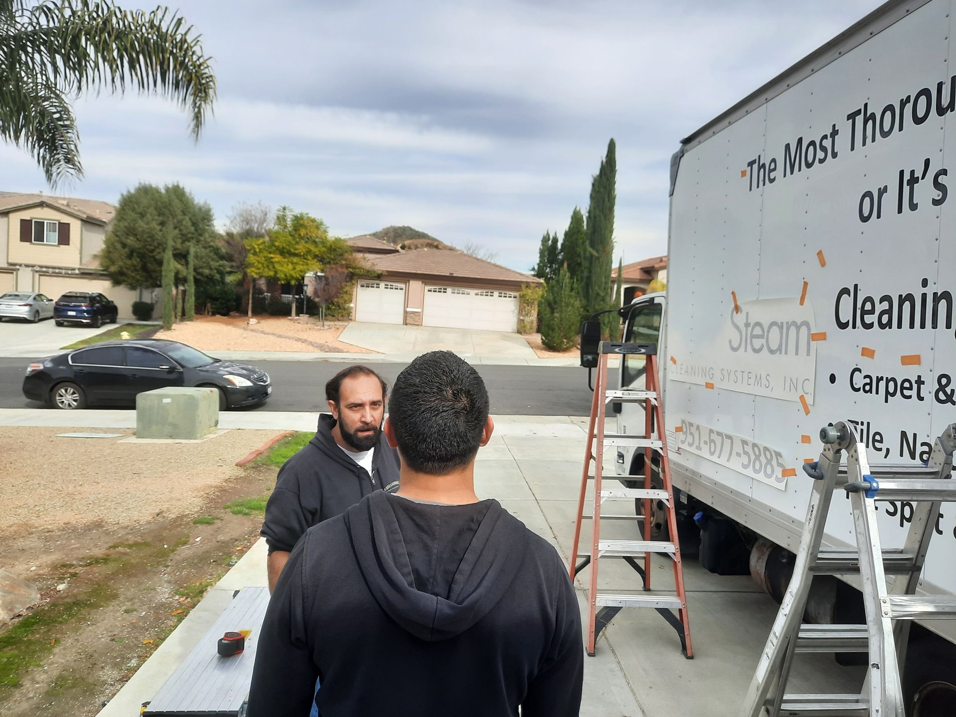 Two men near a Steam Cleaning truck and ladder on a suburban street