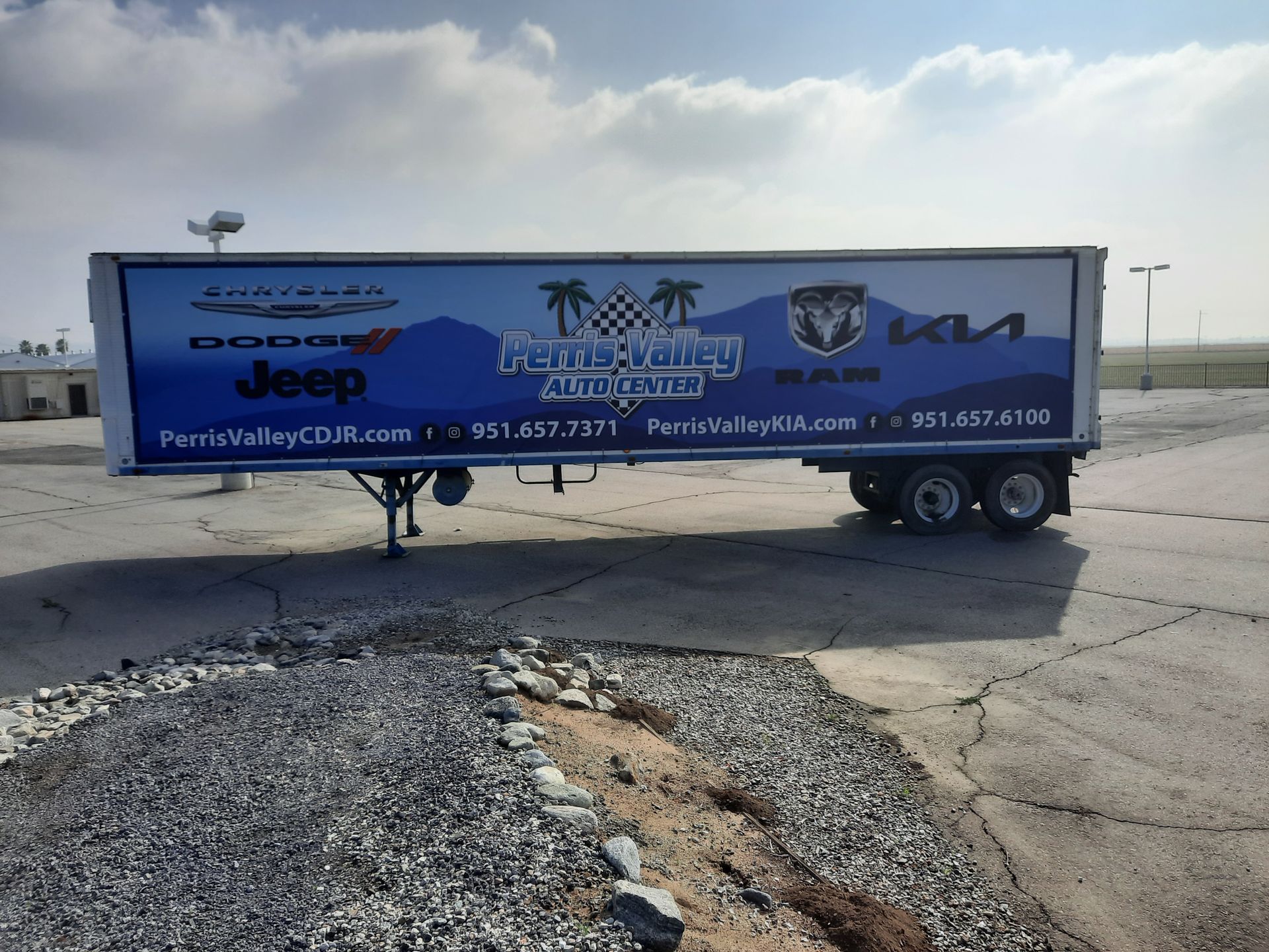 Blue billboard trailer parked on gravel under a cloudy sky, advertising local businesses.