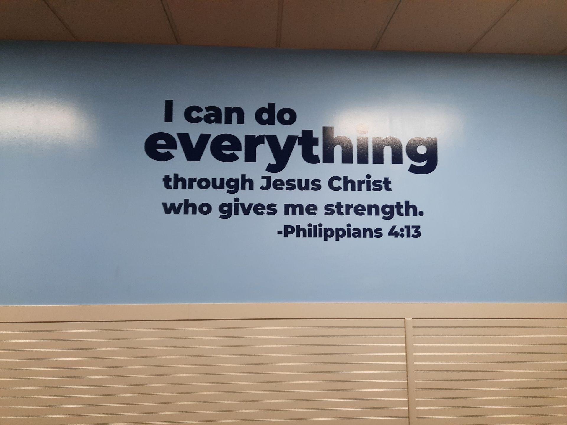 Wall sign with Philippians 4:13 on a blue wall above light wood paneling.