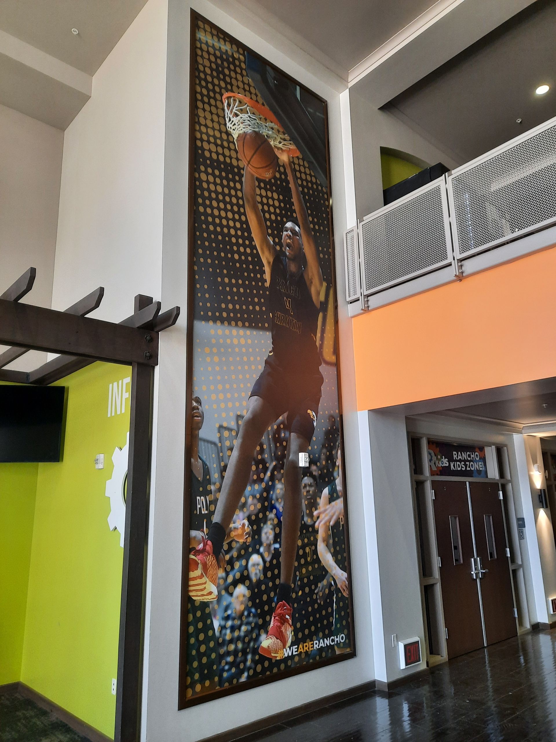 Large indoor mural of a basketball player on a lobby wall beside a mezzanine with orange and white railings.