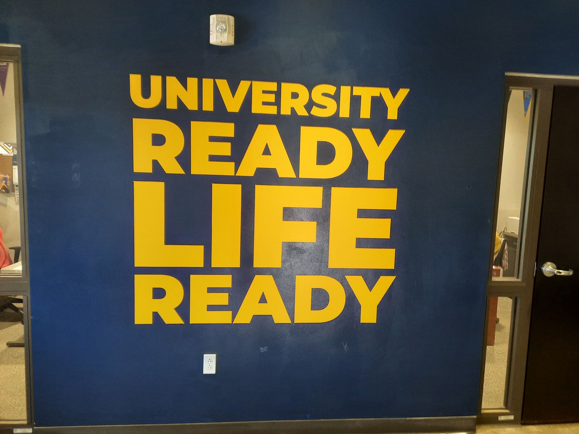 Blue wall mural with yellow text reading “University Ready Life Ready” in a hallway