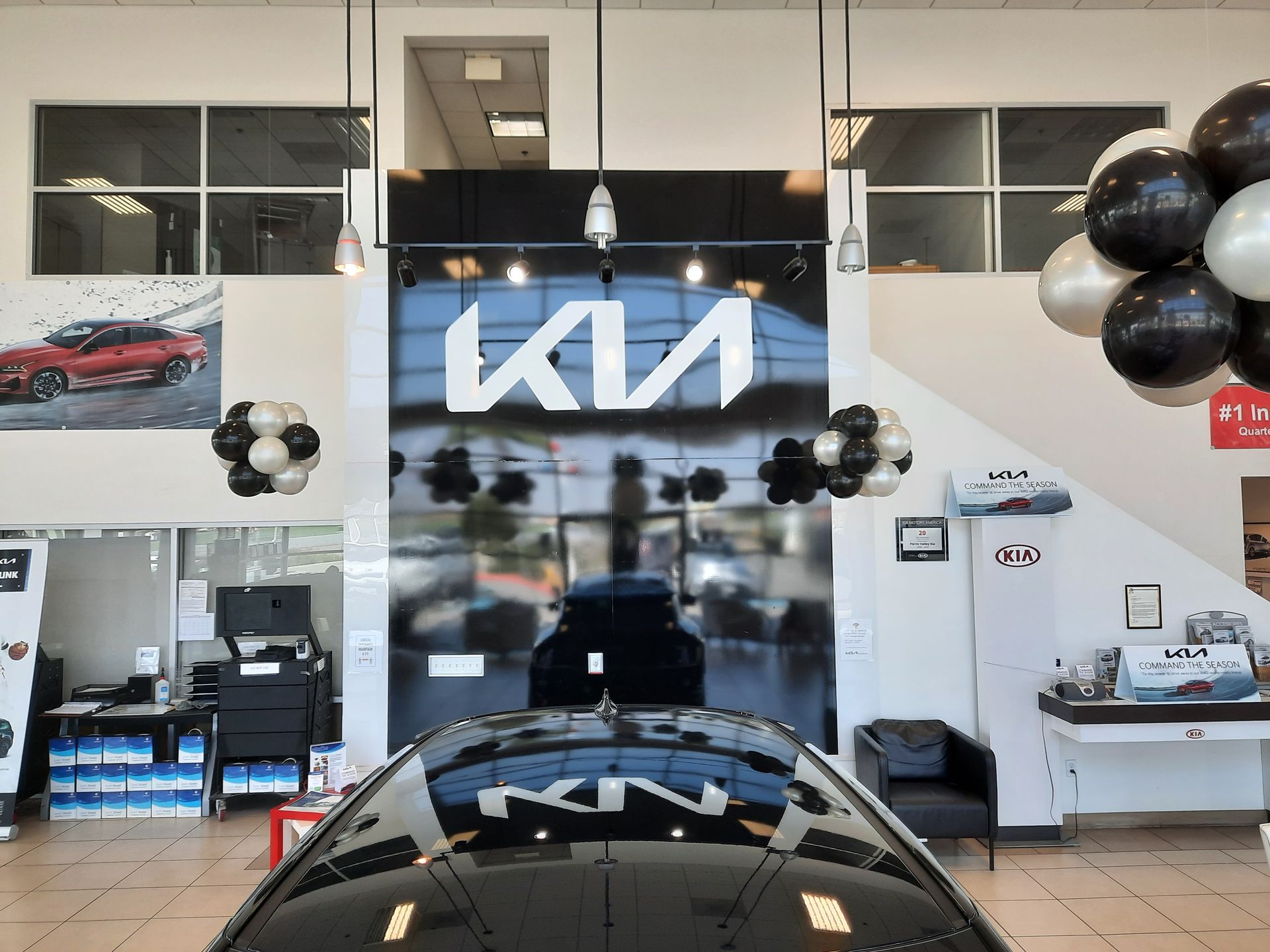 Kia dealership showroom with black and silver balloons, red car poster, and a car front in foreground