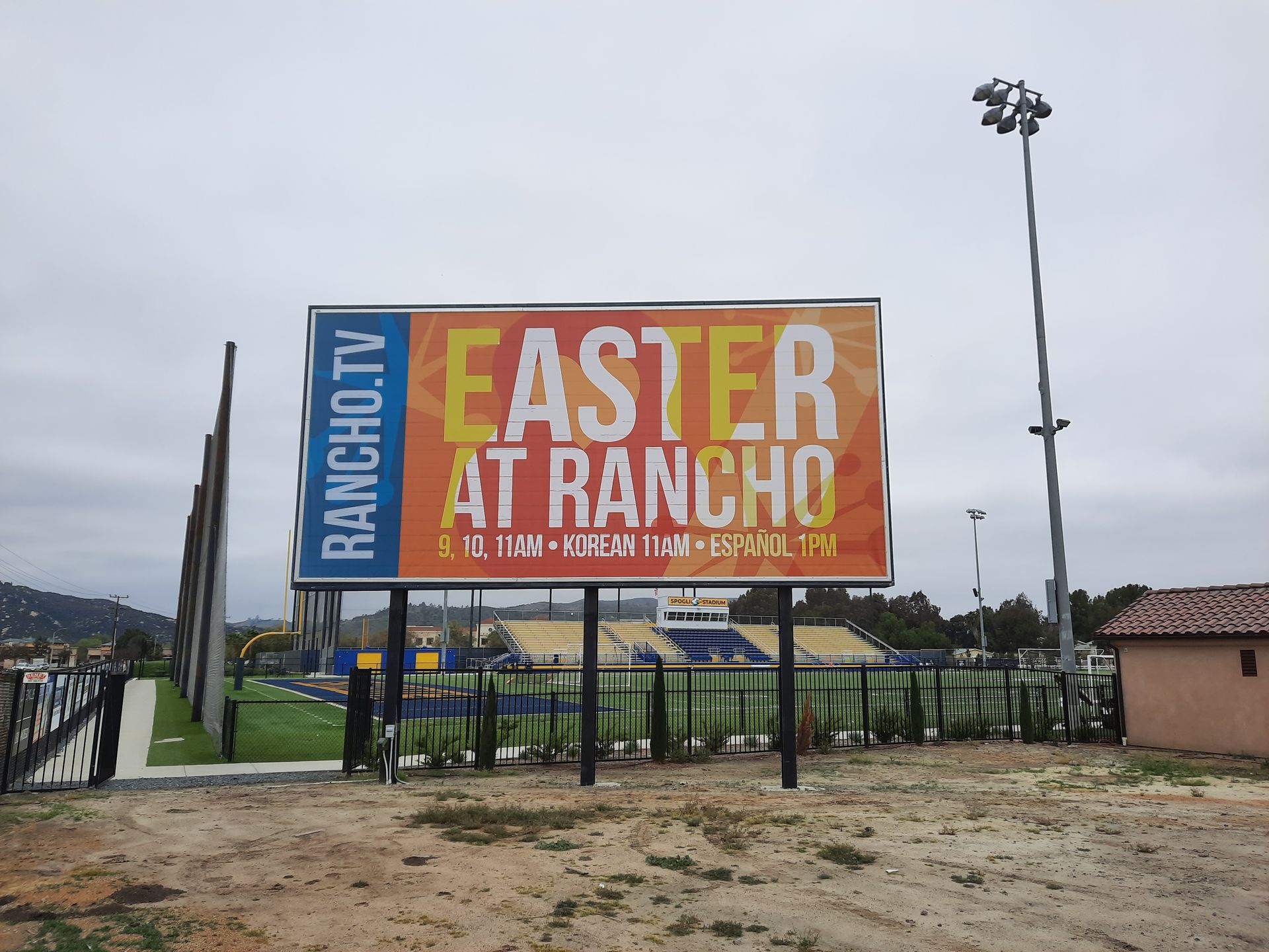 Large orange “Easter at Rancho” event banner on a fenced field under a cloudy sky.