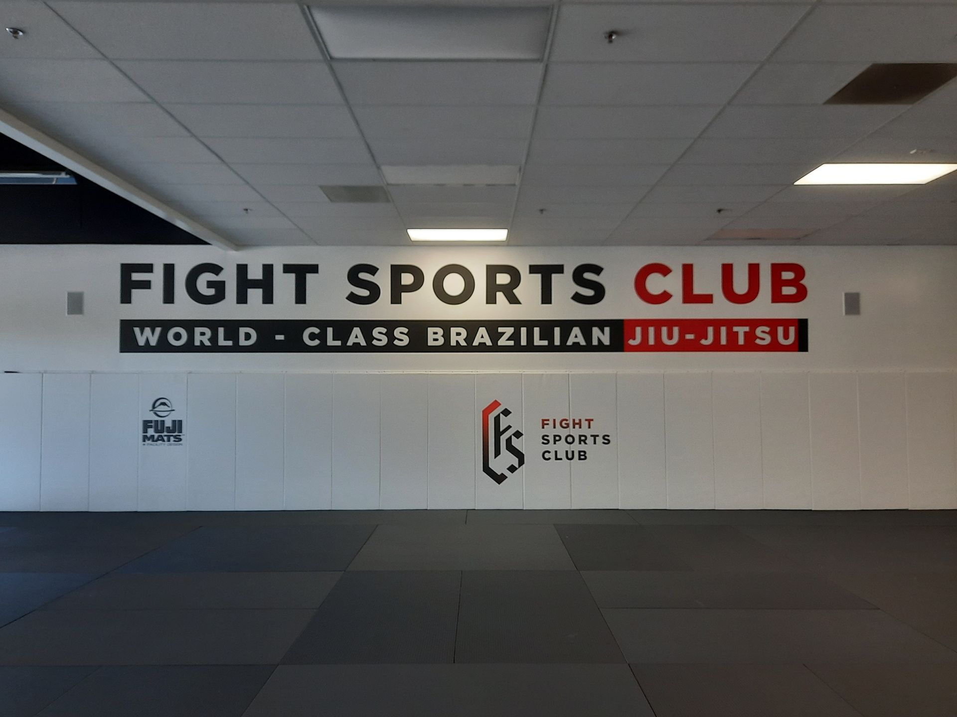Empty martial arts studio with mats and wall sign reading “Fight Sports Club” and “World-Class Brazilian Jiu-Jitsu”