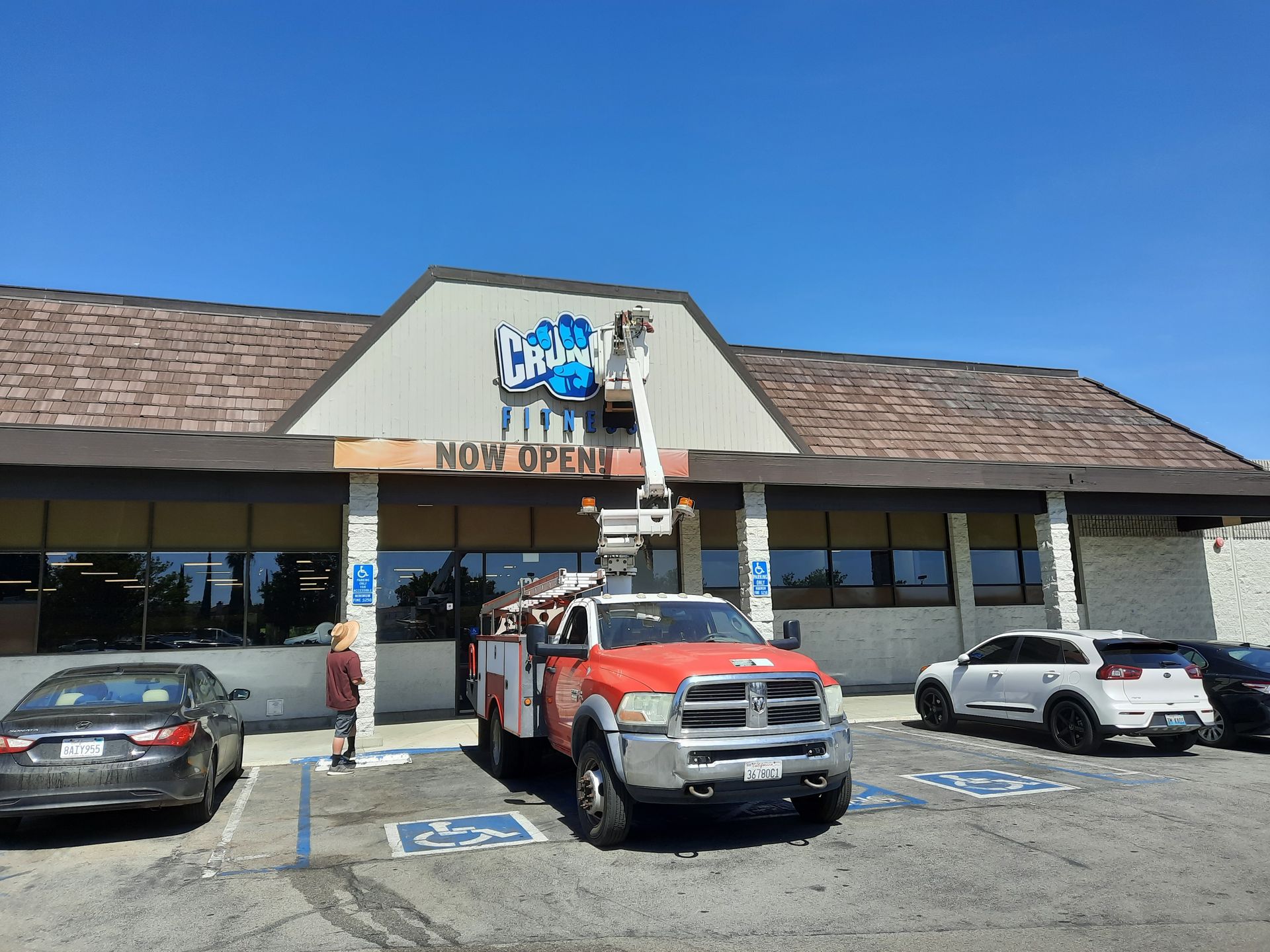 Strip mall storefront with a “NOW OPEN” sign and several parked cars, including a red pickup in front.