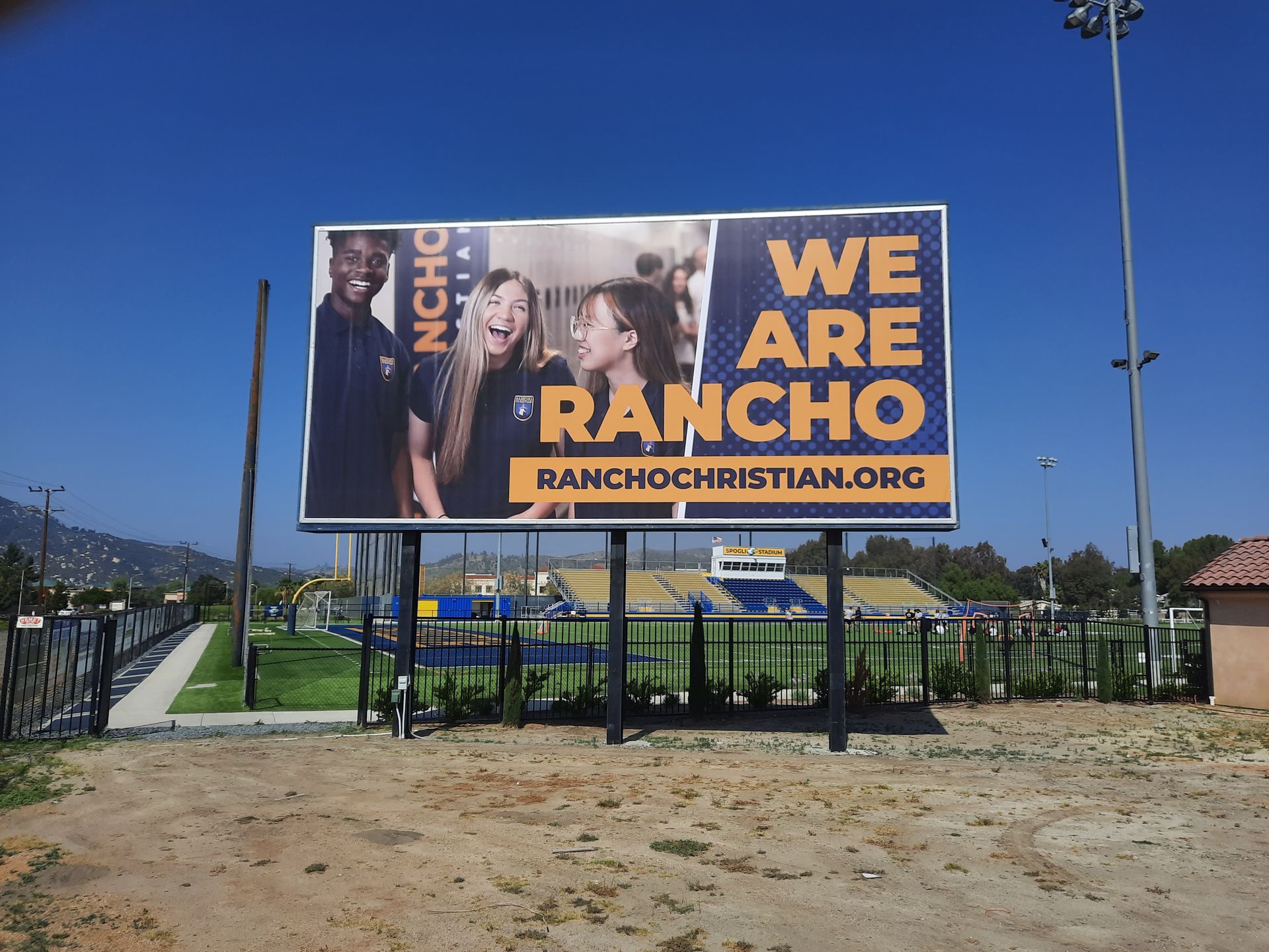 Billboard for Rancho Christian Church reading “WE ARE RANCHO” beside a road and fence