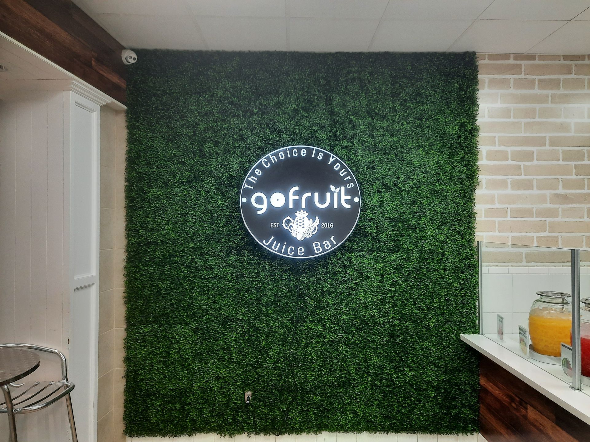 Green faux-hedge wall with a round “gofruit” café sign inside a juice bar.