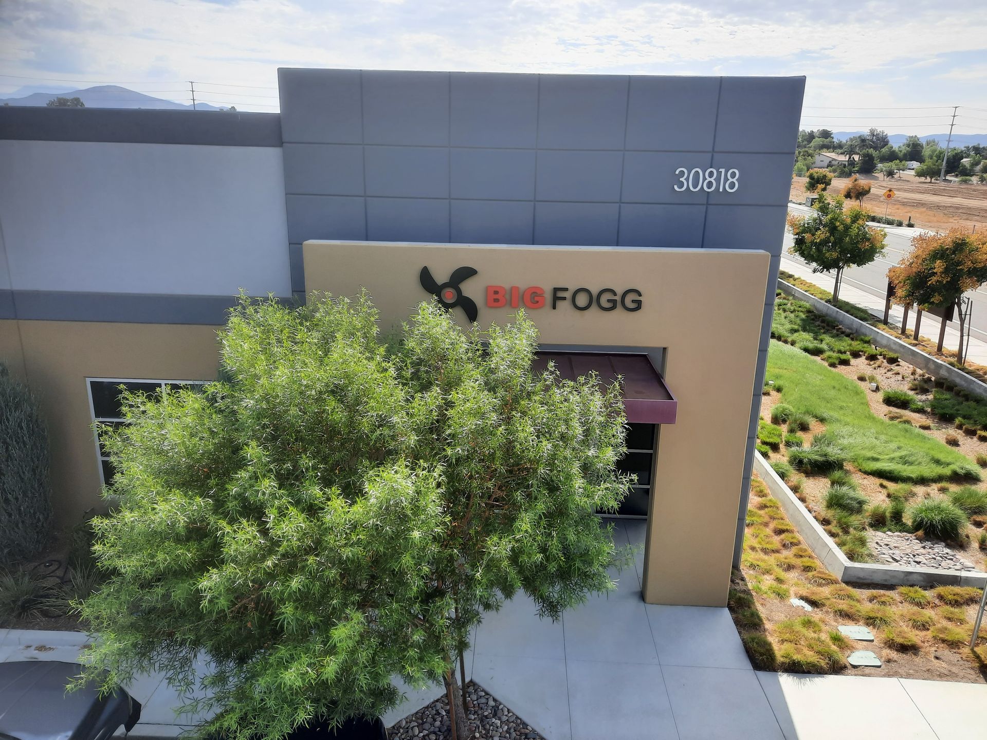 Entrance to a BE FOGG store with a tree partly blocking the view, beside a landscaped walkway