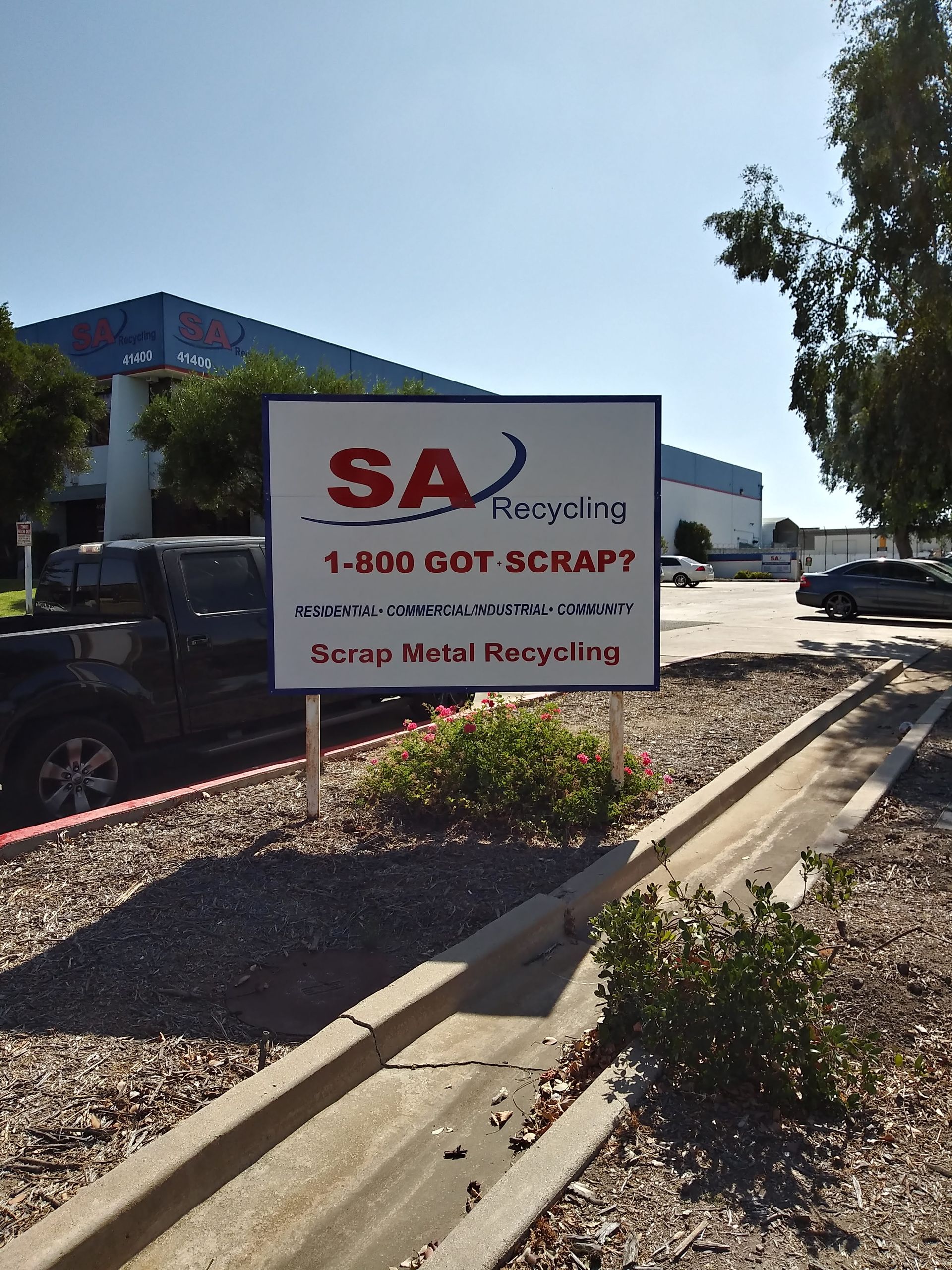 White sign for SA Recycling beside a curb in a sunny parking lot, with a black truck nearby.